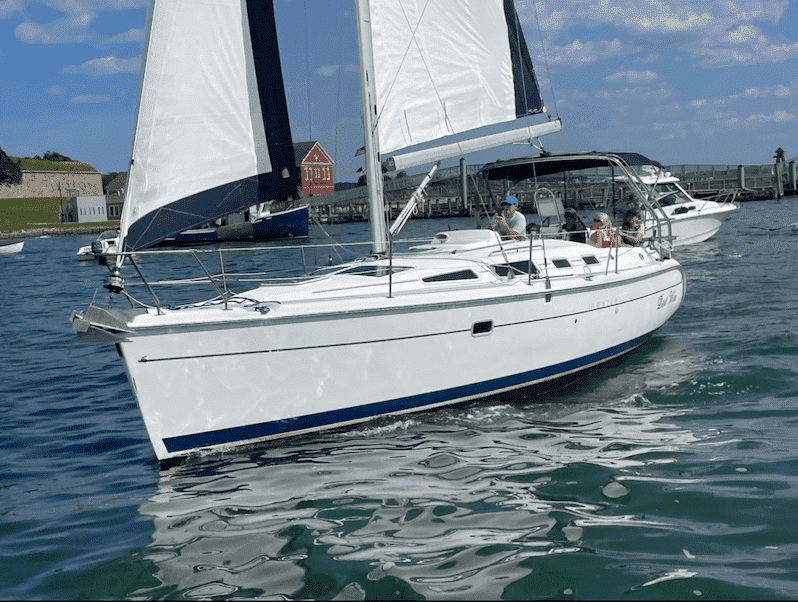 White sailboat cruising on calm water near a harbor, with people onboard enjoying a sunny day.