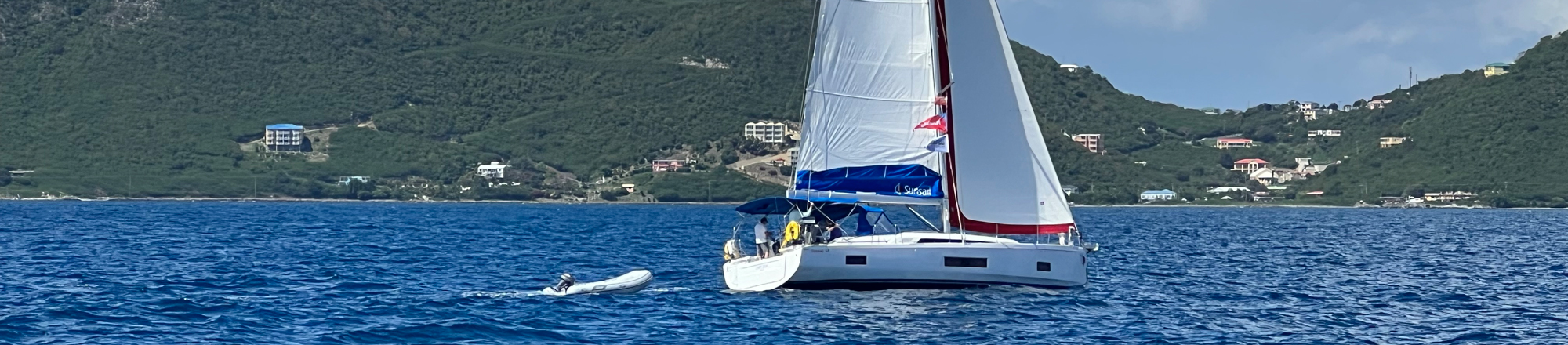 Sailboat about to gybe - cruising near lush island shoreline, vibrant blue sea and sky, ideal for sailing adventures.
