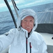 Man in white sailing jacket smiles on deck with ocean backdrop, showcasing adventure and marine technology.