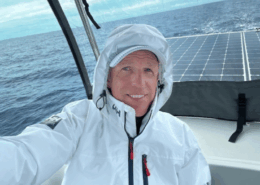 Man in white sailing jacket smiles on deck with ocean backdrop, showcasing adventure and marine technology.