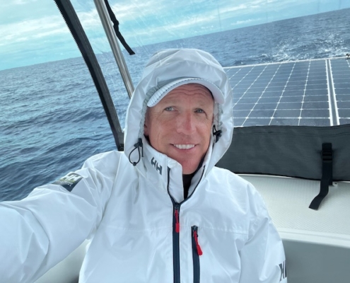 Man in a white jacket smiles on a sailboat with solar panels and ocean backdrop.