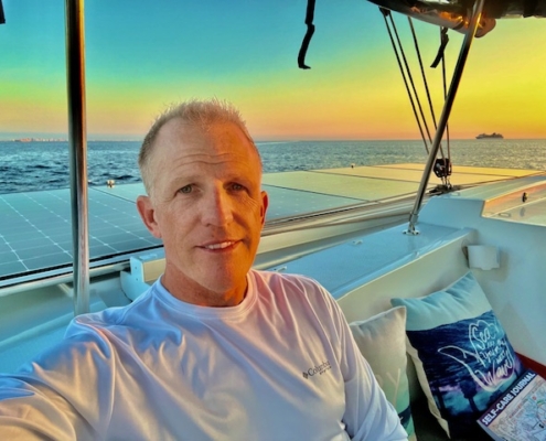 Man enjoying a sunset view from a sailboat deck, with ocean horizon and colorful sky in the background.