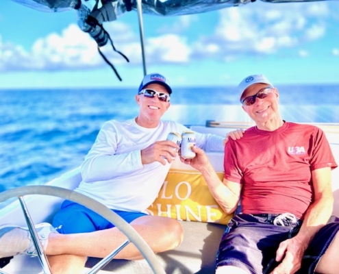 Two friends relaxing and toasting on a sailboat under a sunny sky, enjoying a perfect day at sea.