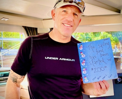 Smiling man on a boat holding a note about starting a sailing fund with $200, inside a sunlit cabin.