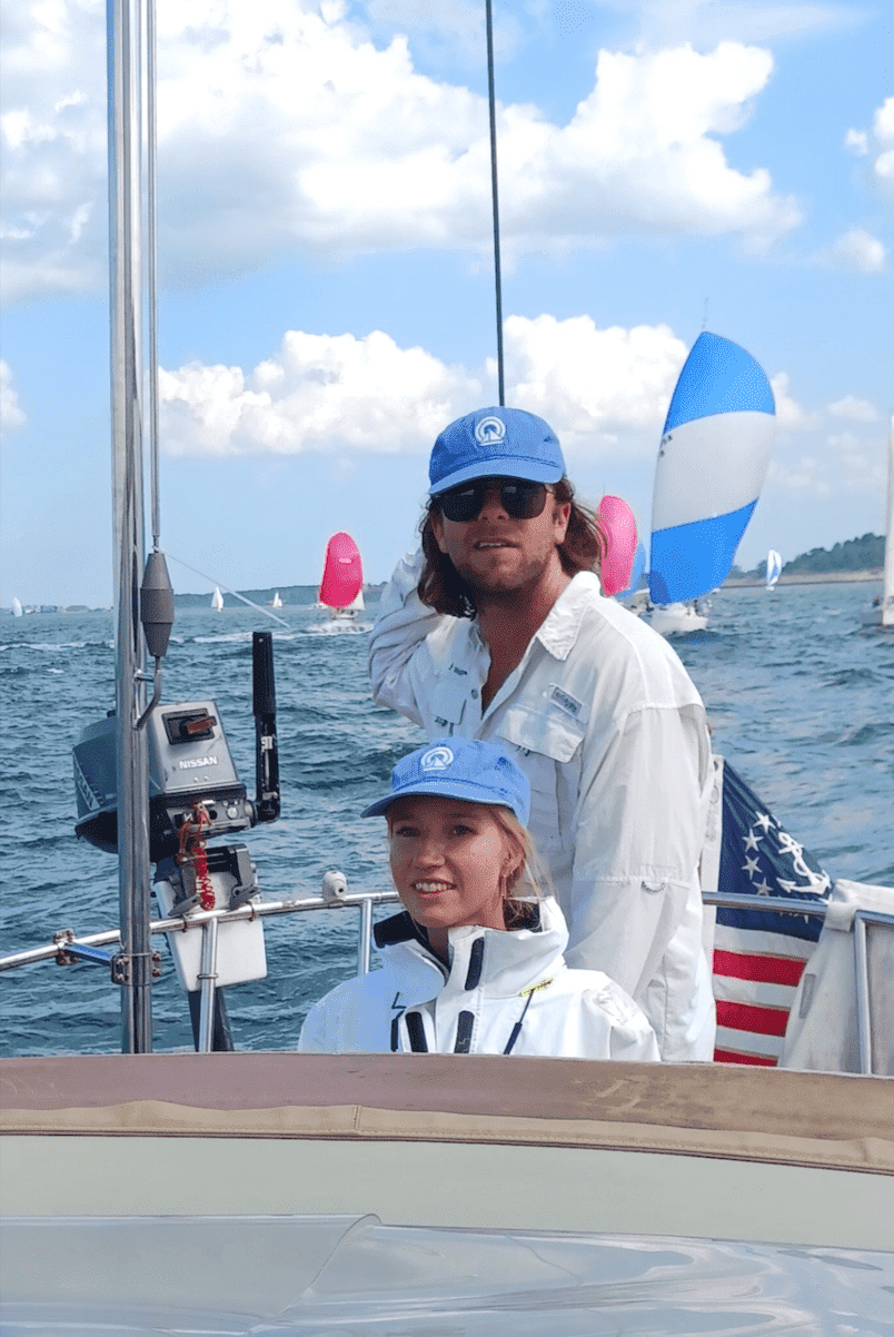 Two sailors on a boat enjoy a sunny day on the water, surrounded by colorful sails and a clear blue sky.
