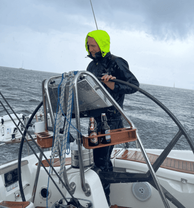 Man in rain gear steering sailboat in stormy weather with drink bottles on helm, showcasing adventurous sailing.