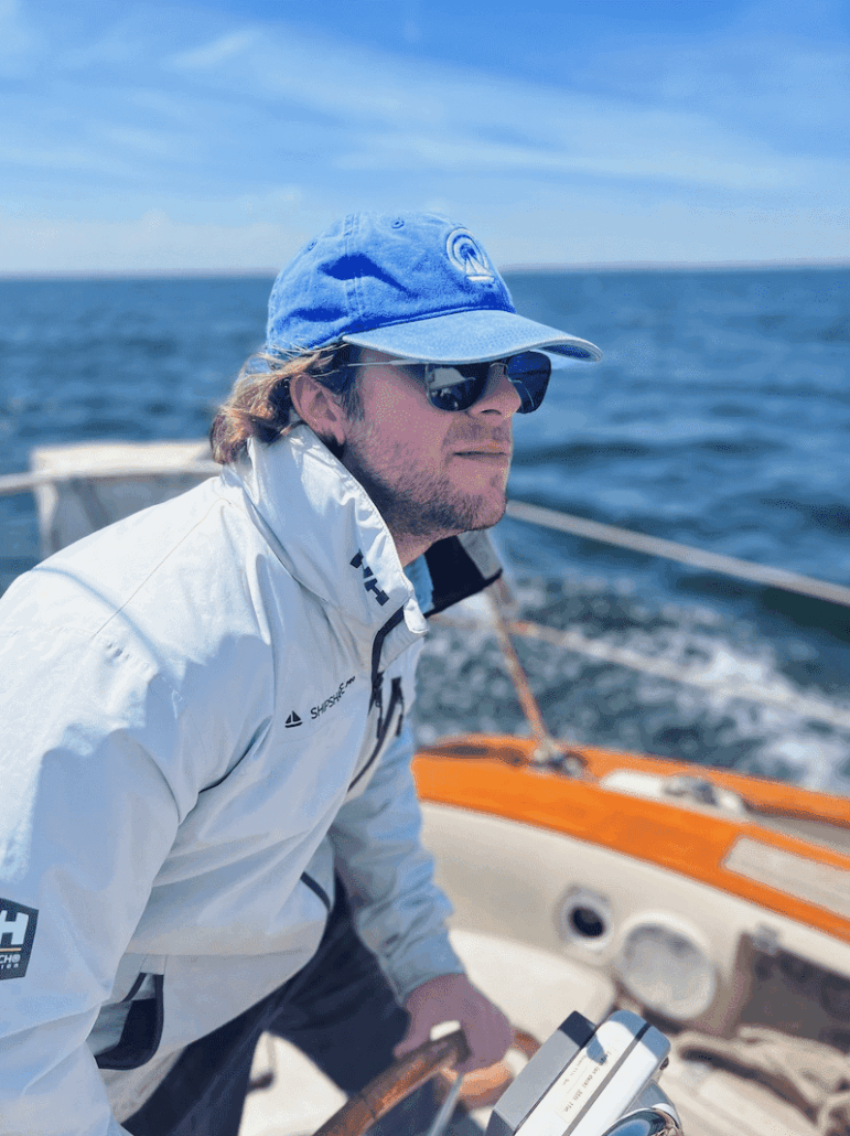 Person sailing a boat on open water, wearing a blue cap and white jacket, under a clear blue sky.