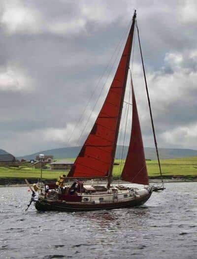Sailing yacht with red sails gliding on scenic waters near green coastal hills under a cloudy sky.