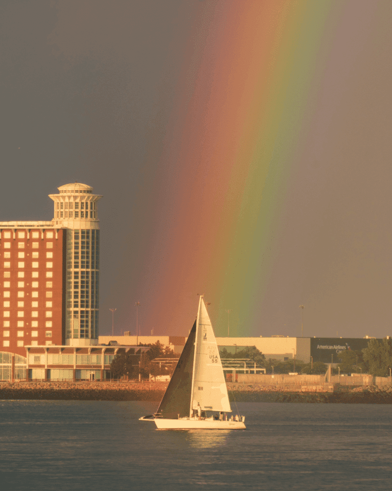 Sailboat glides on water near buildings with a vibrant rainbow in the background.