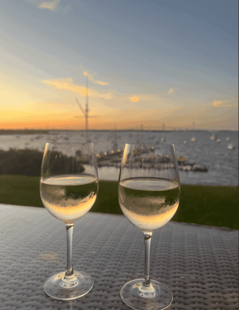 Two glasses of white wine on a wicker table overlook a serene sunset at a marina, with yachts anchored in the harbor.