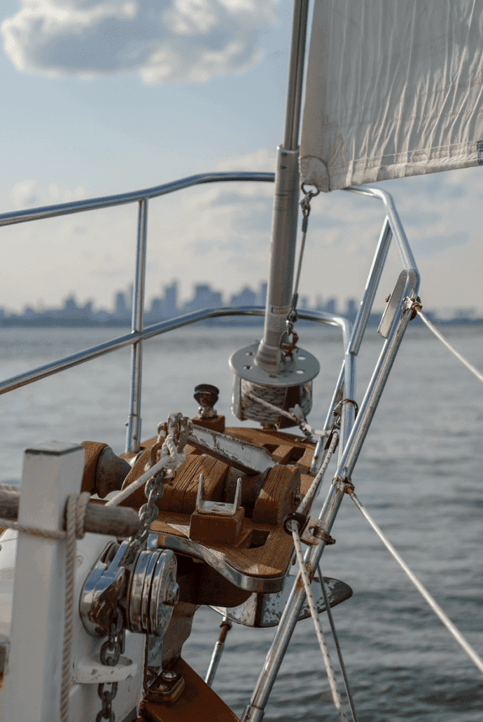 Sailing ship's bow with ropes and winch, calm sea, and distant city skyline under a blue sky.