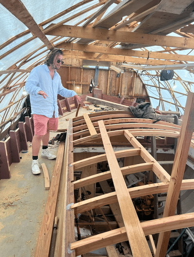 Man inspects detailed wooden sailboat construction inside a shipyard, surrounded by beams and tools.