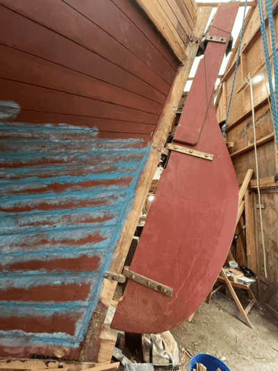 Wooden sailboat rudder under repair in a workshop, showcasing detailed craftsmanship and marine hardware.