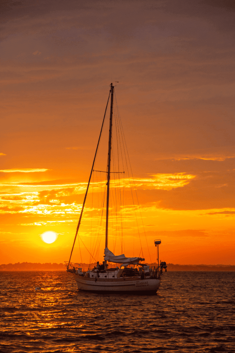 Sailboat cruising on tranquil sea at golden sunset, with vibrant sky and sun on the horizon.
