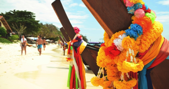 Colorful floral garlands adorn longtail boats on a sandy beach, with tourists enjoying the sunny day.