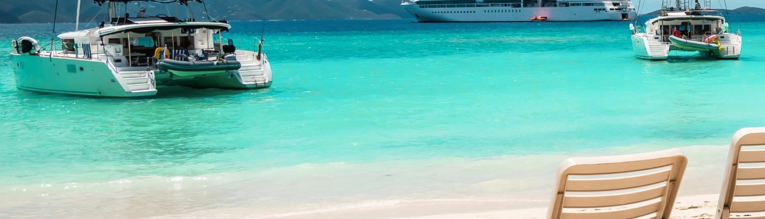 Tropical beach with sailboats and cruise ship on turquoise water, under a clear sky, viewed from sandy shore with chairs.