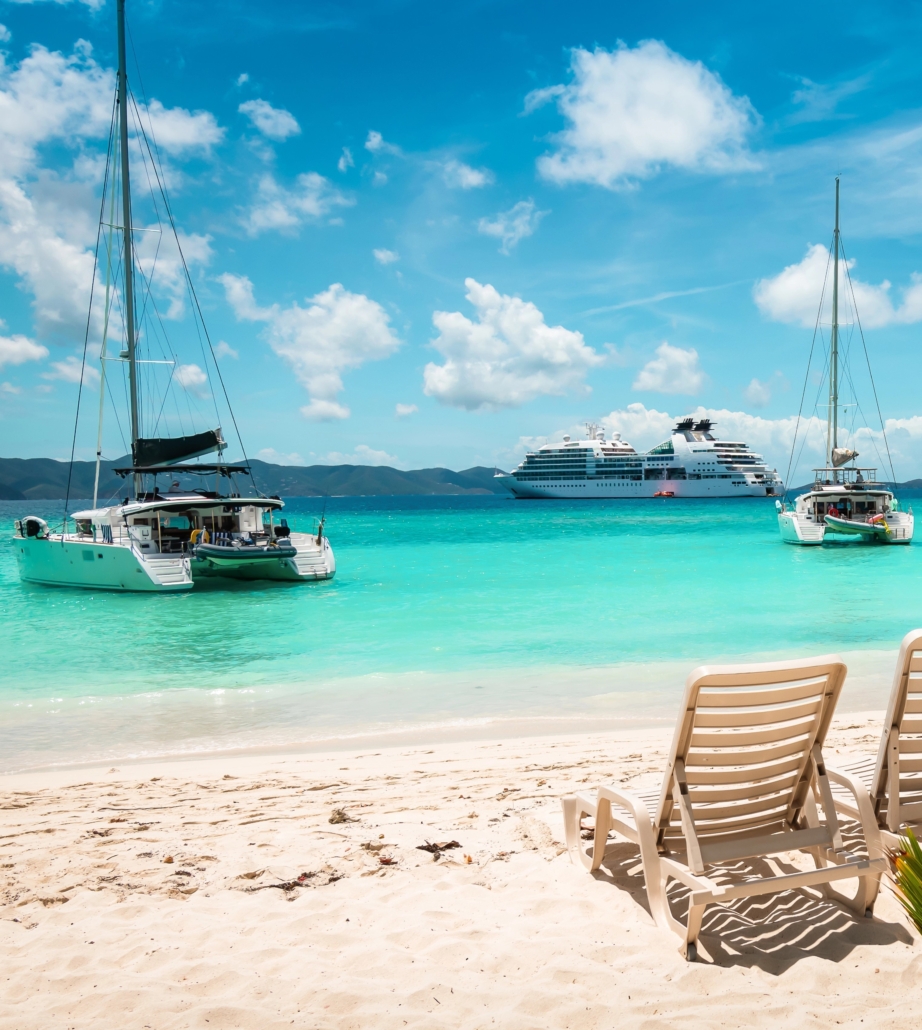 Tropical beach with sailboats and a cruise ship in turquoise waters, two lounge chairs on sandy shore under blue sky.