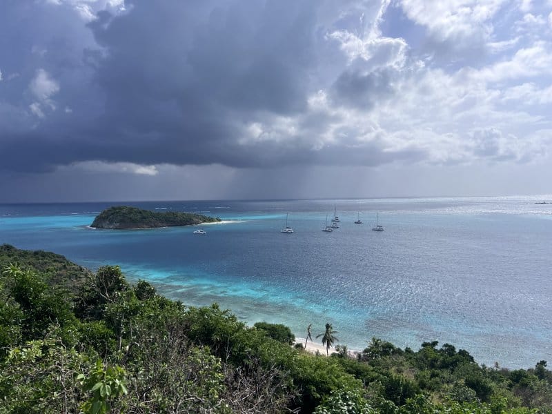 Sailboats anchored near a tropical island with turquoise waters under a dramatic cloudy sky.