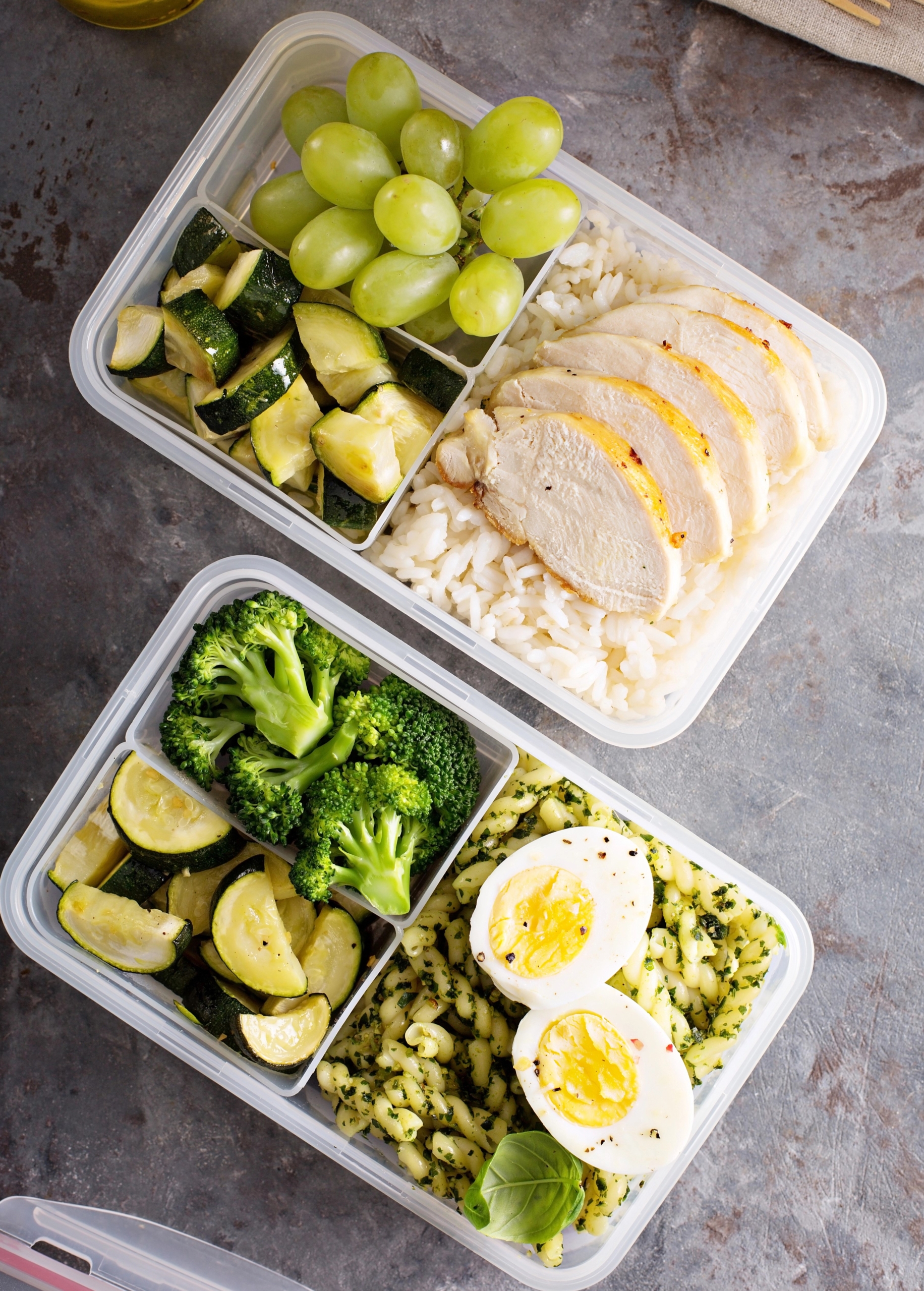 Healthy meal prep with chicken, rice, grapes, broccoli, zucchini, pasta, and eggs in divided containers on a countertop.
