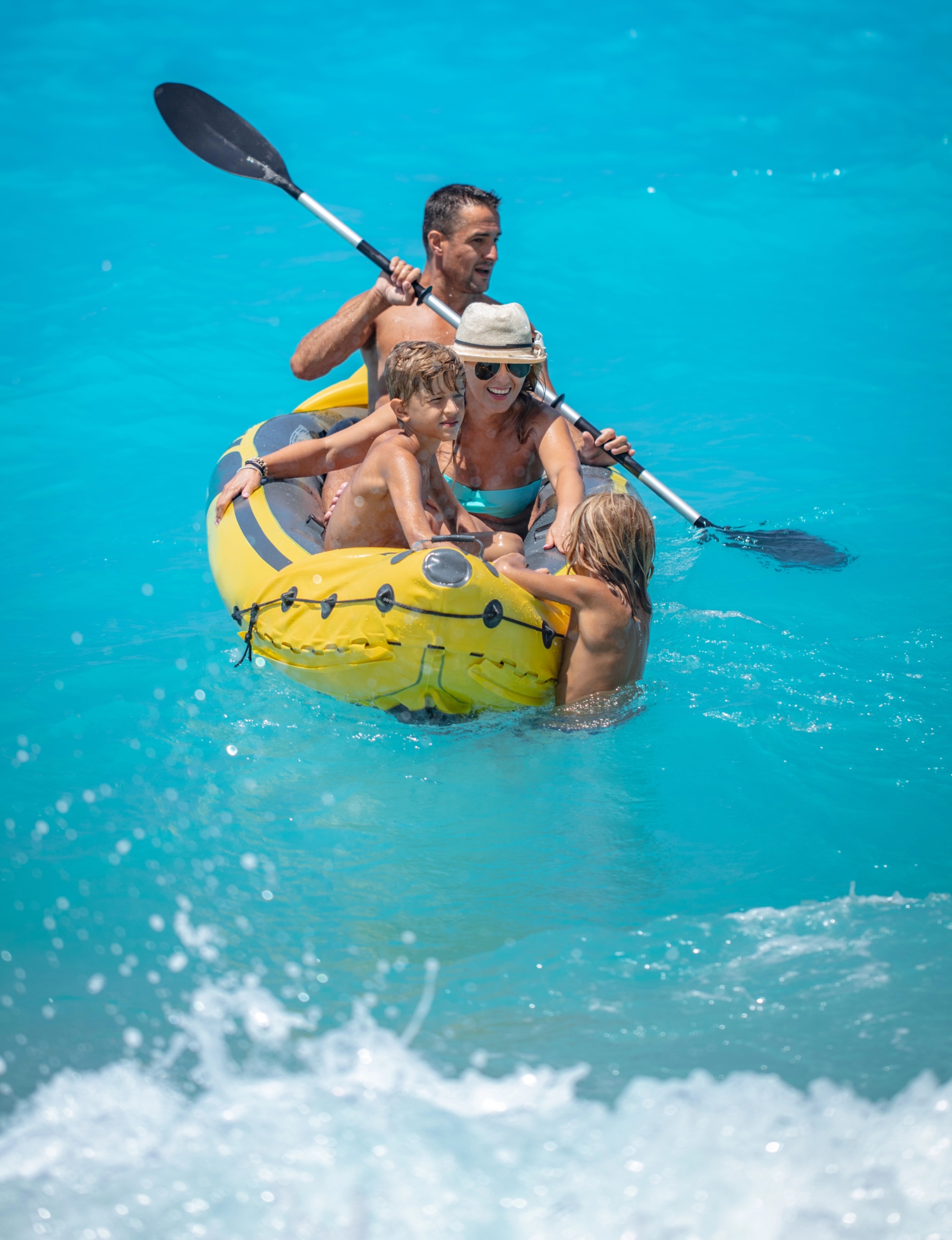 Family enjoying a sunny day kayaking on clear blue ocean waters, paddling together in a yellow inflatable kayak.