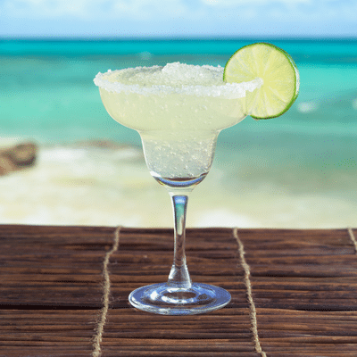 Chilled margarita with lime on a wooden table, overlooking a tropical beach and turquoise sea under a clear sky.