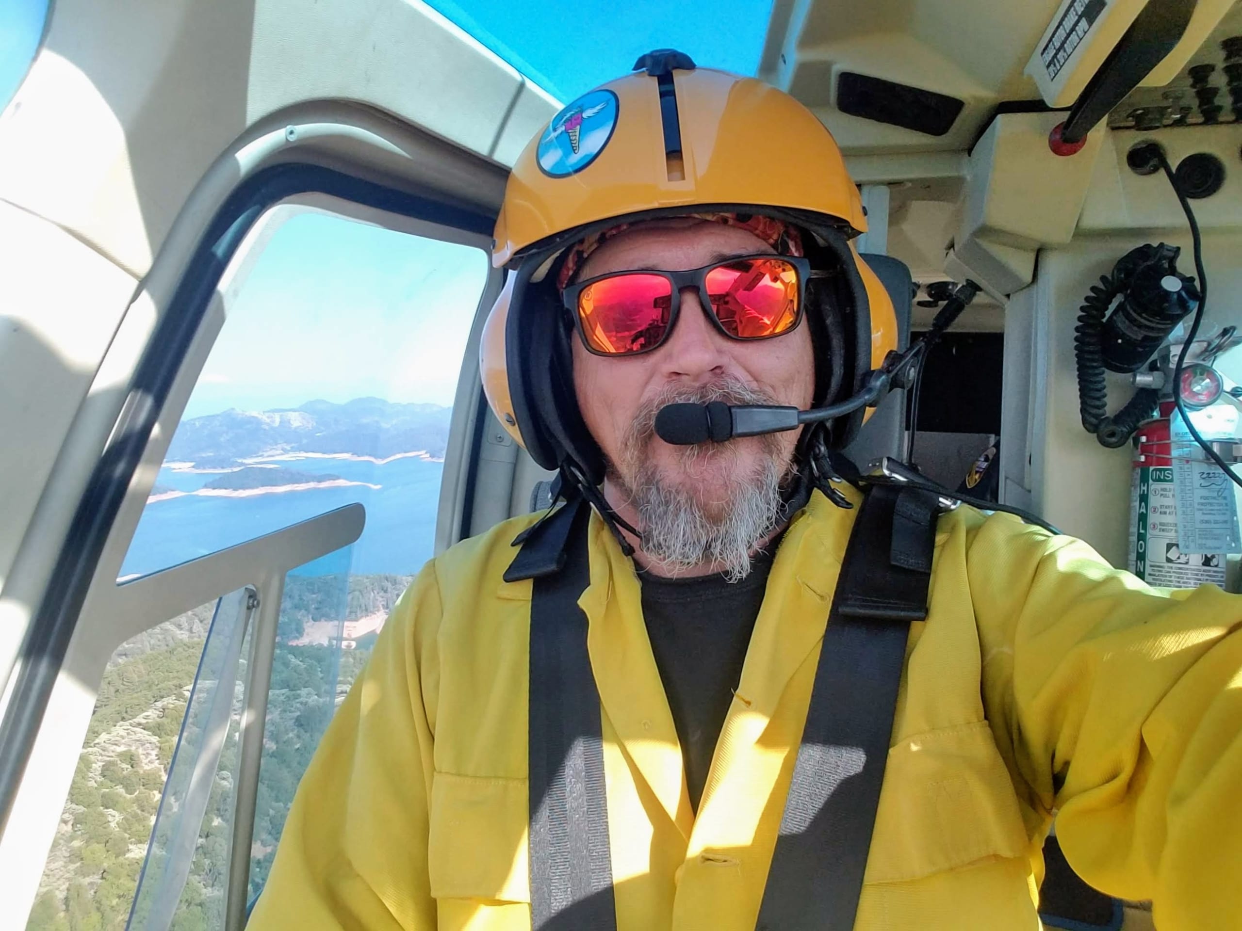 Pilot in yellow gear flying over scenic coastline, with ocean and islands visible through helicopter window.
