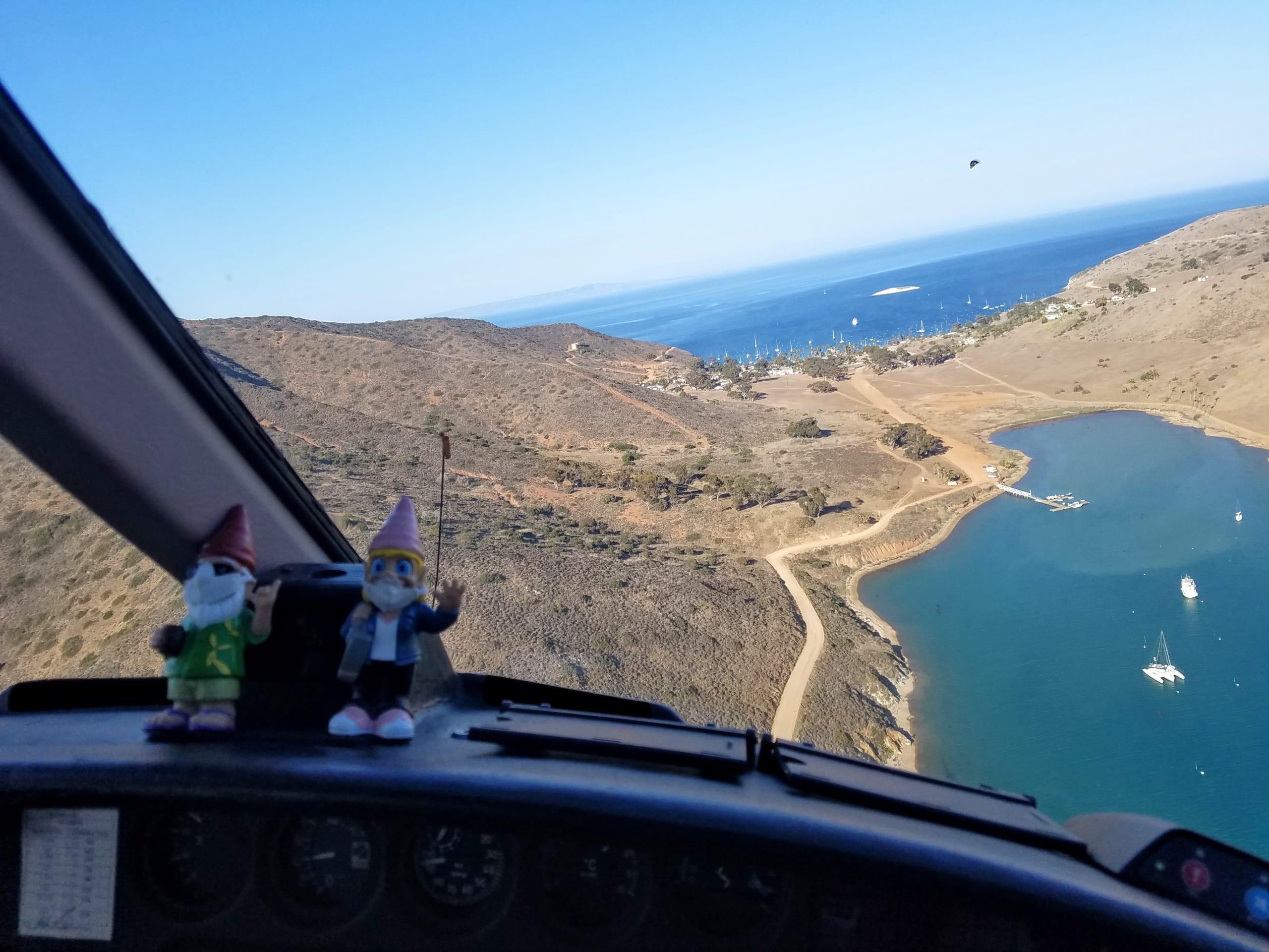 Aerial view of coastline with sailboats in harbor, seen from cockpit with two gnome figures on dashboard.