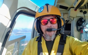 Capt. Gordy Cox in yellow safety gear and sunglasses flying a helicopter, overlooking a scenic coastline, after performing a proper risk assessment