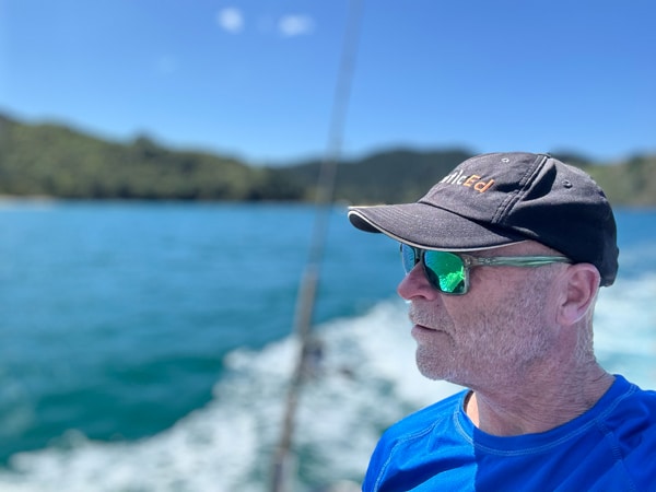 Grant Headifen enjoying sailing, wearing sunglasses and cap, gazing at sea; clear blue sky and water in the background.