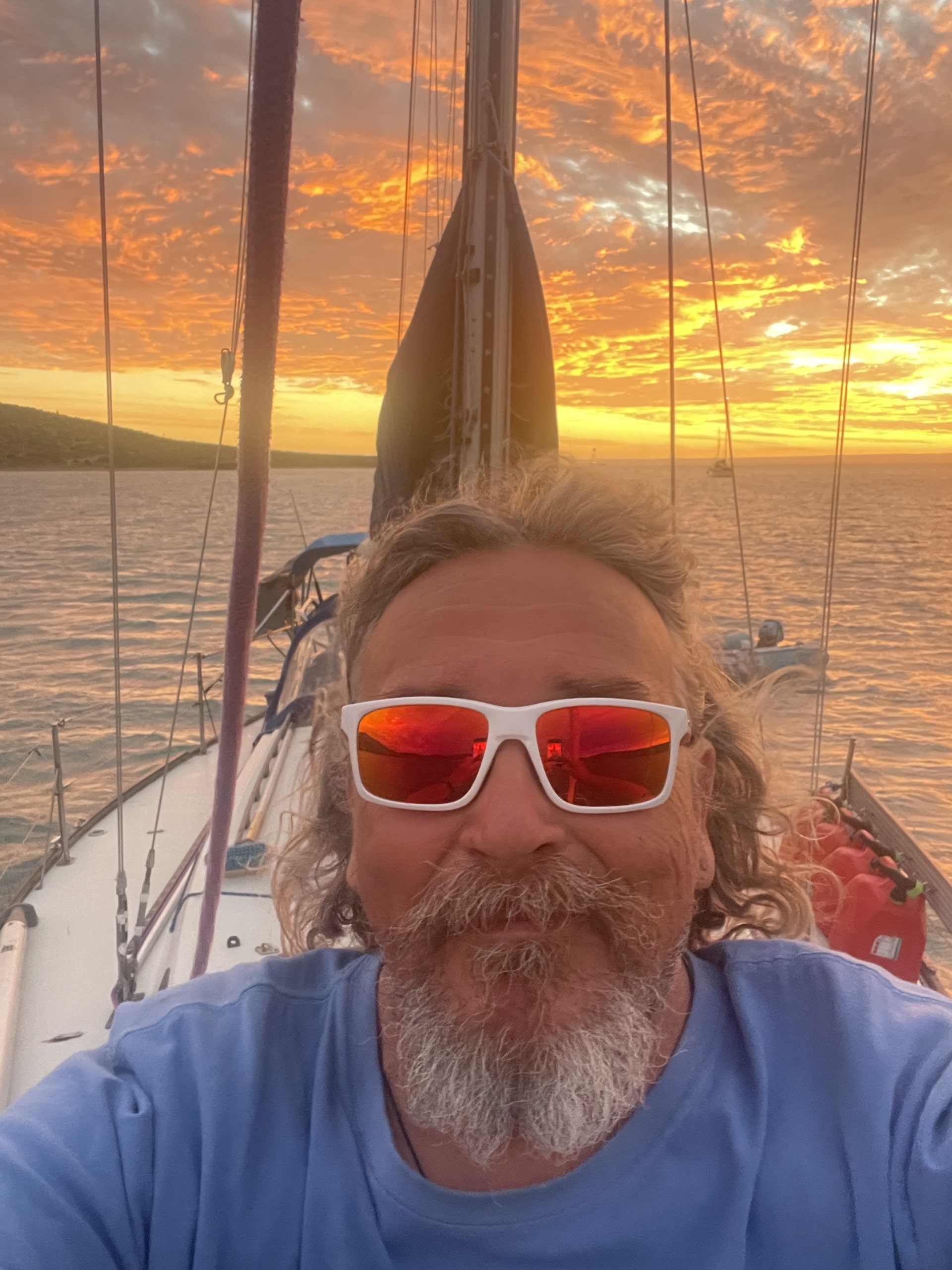 Happy sailor with sunglasses enjoys a stunning sunset on a sailboat, sailing into an orange-lit horizon.