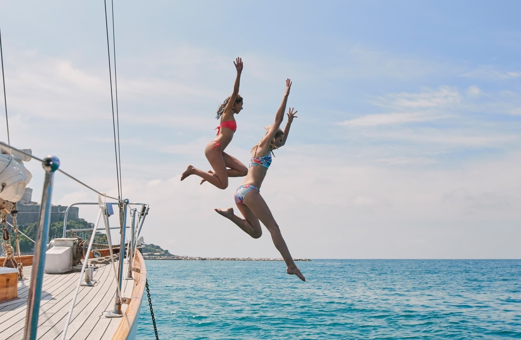 Two women in bikinis joyfully leap off a sailboat into the clear blue ocean on a sunny day.