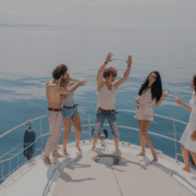 Group of friends dancing on yacht deck, enjoying a sunny day at sea with drinks, laughter, and carefree fun on a luxury crewed charter