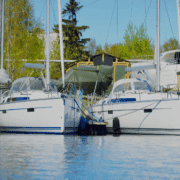Two sailboats docked side by side in a tranquil marina, surrounded by lush trees and clear skies.