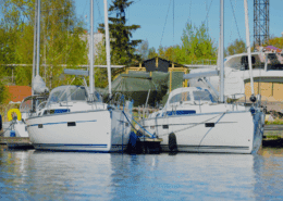 Two sailboats docked side by side in a tranquil marina, surrounded by lush trees and clear skies.