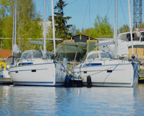 Two sailboats docked side by side in a tranquil marina, surrounded by lush trees and clear skies.