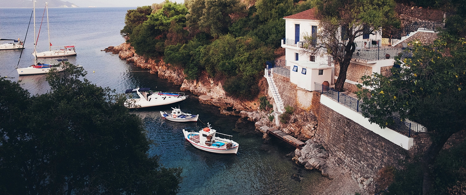 Secluded coastal house with moored boats and sailboats in a serene bay surrounded by lush greenery.