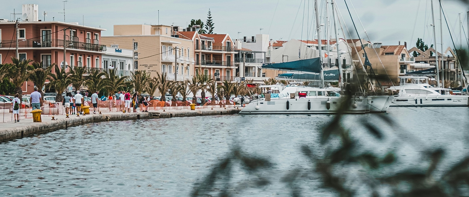 Sailboat docked at a picturesque harbor with people strolling along a lively waterfront promenade.
