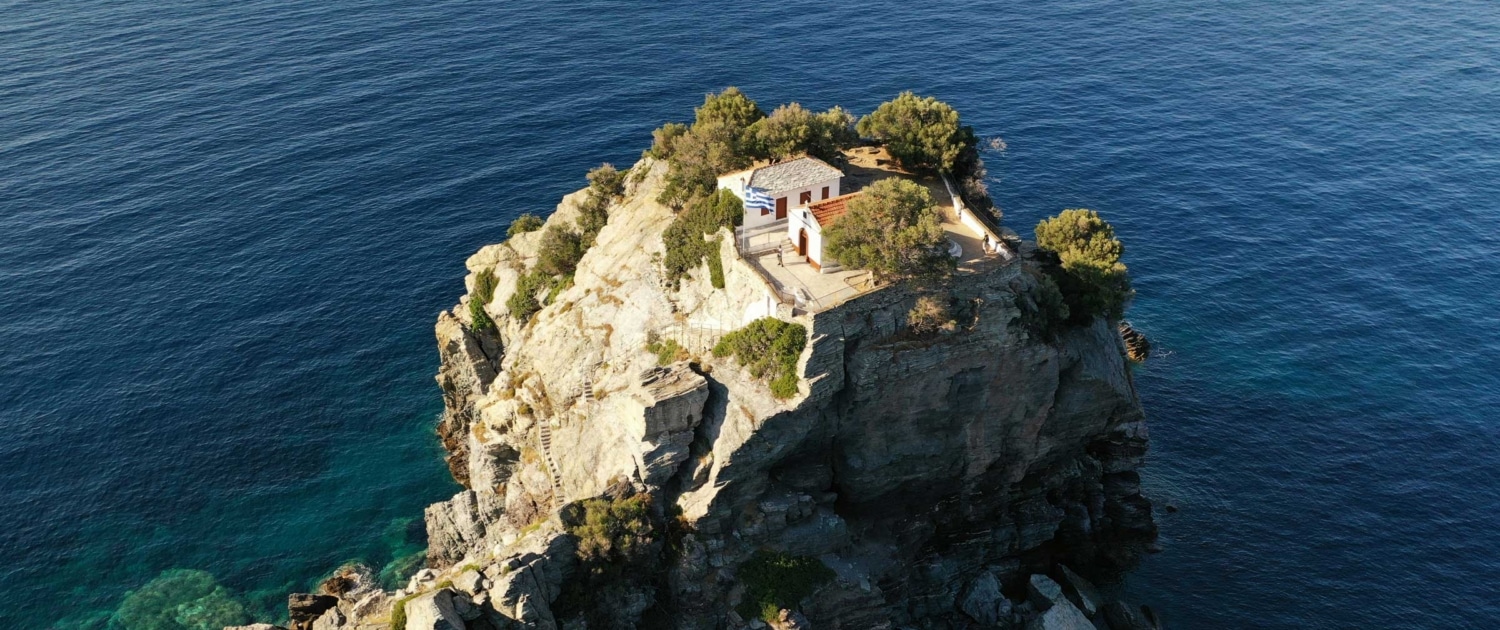 Secluded coastal chapel on a rocky cliff overlooking the deep blue sea, surrounded by lush greenery.
