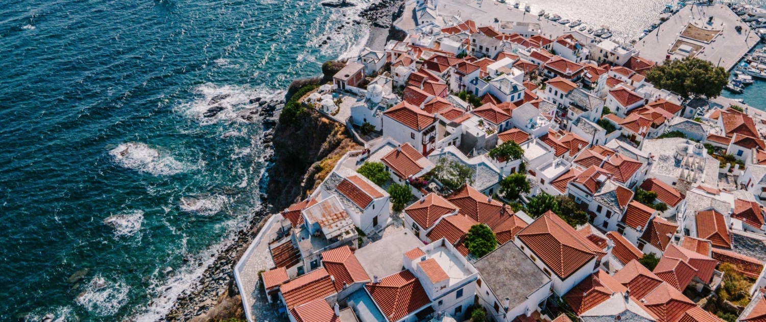 Aerial view of a coastal Mediterranean village with white houses and red roofs by the sea, near a marina.