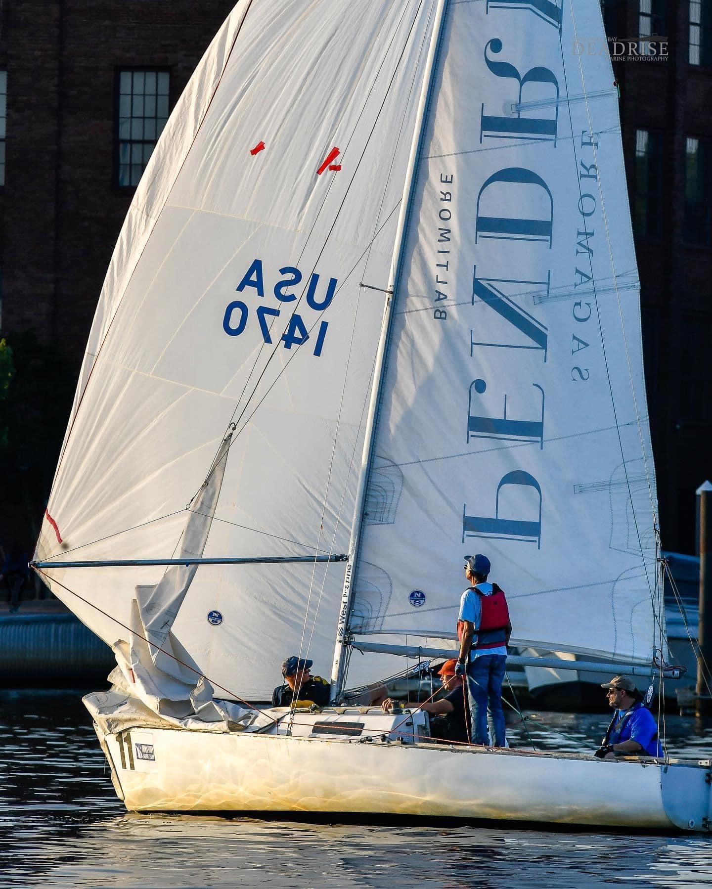 Sailboat with crew navigating calm waters near a cityscape, featuring crisp white sails and a vibrant sunset glow.