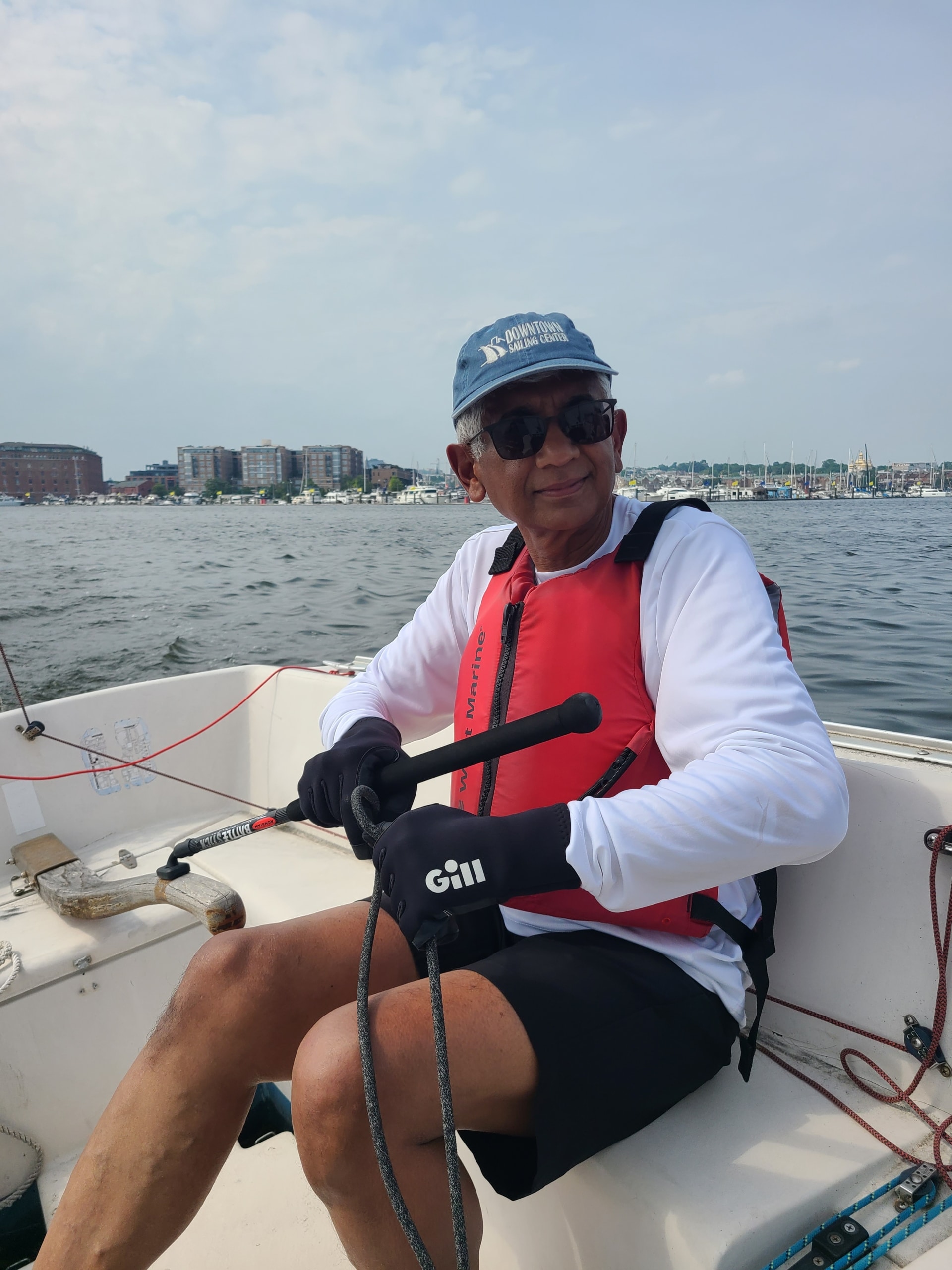 Man sailing on calm waters with city skyline in background, wearing red life jacket and blue hat, enjoying a sunny day.