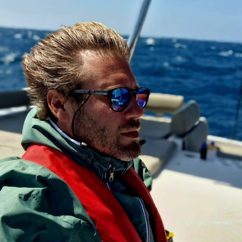 Man in sunglasses and red life jacket sailing on the ocean with blue skies in the background.