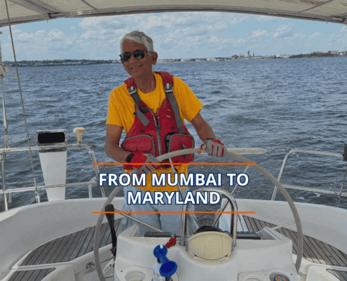 Person sailing with a smile under sunny skies, wearing a red vest and yellow shirt, captioned From Mumbai to Maryland.