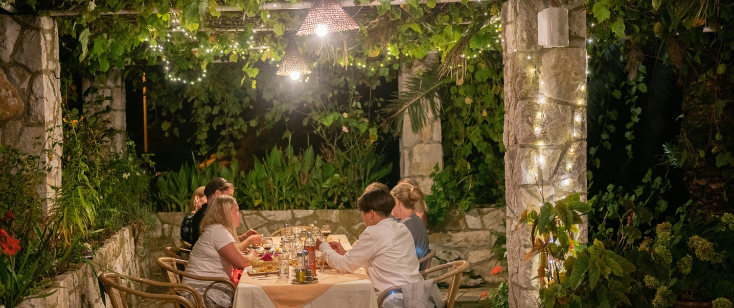 Outdoor dinner under vine pergola with friends, ambient lights, and lush greenery creating a cozy evening atmosphere.