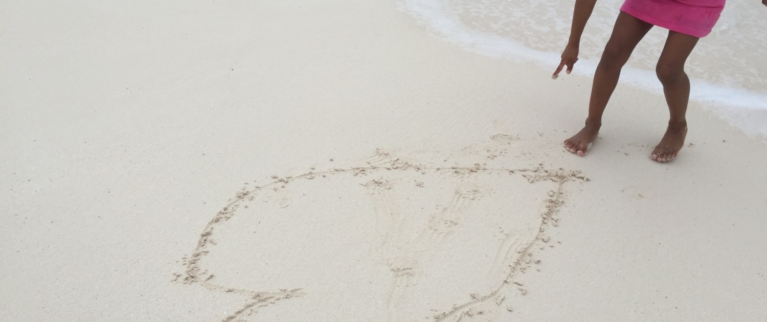 Child in pink dress drawing a heart in the sand on a beach by the sea.