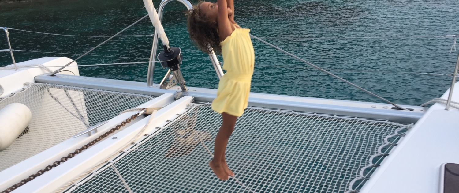 Child joyfully leaps on a catamaran's netting, surrounded by serene blue waters and lush greenery.