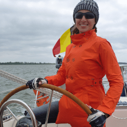 Smiling sailor in an orange jacket steering a boat on a cloudy day, with a colorful flag in the background.