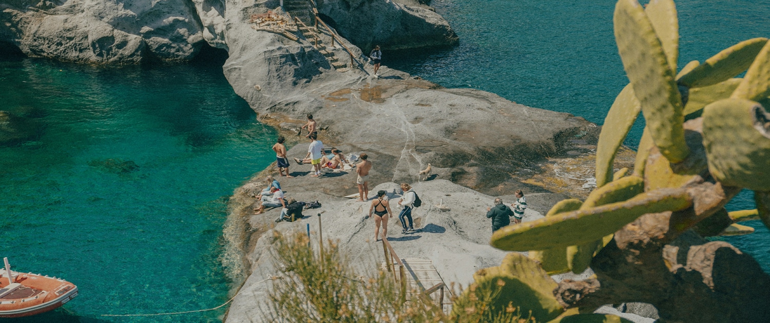 People relaxing on rocky cliffs by turquoise sea, near a small boat, with cactus in the foreground.