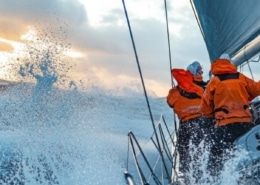Sailors in orange gear navigate a yacht through rough seas, waves splashing, under a cloudy sky at sunset.