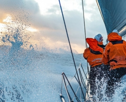 Sailors in orange gear navigate a yacht through rough seas, waves splashing, under a cloudy sky at sunset.